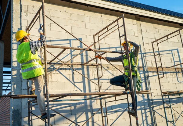 Two construction workers wearing safety gear assemble scaffolding alongside a building, illustrating potential fall hazards and workplace safety concerns under New York labor laws.