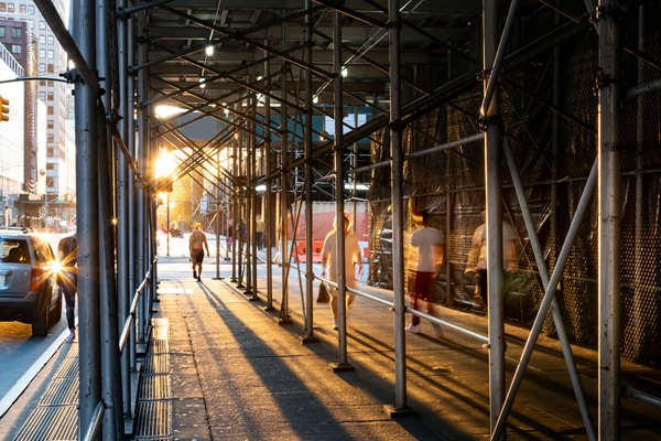 Pedestrians walk through a long sidewalk shed constructed of metal scaffolding pipes along a city street at sunset.