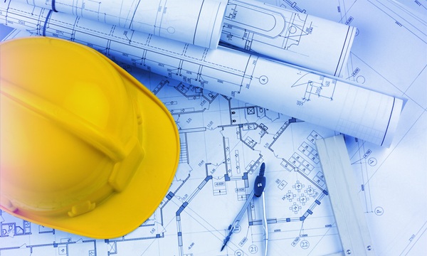 A yellow hard hat rests on a set of architectural blueprints and rolled construction plans for an NYC project site, alongside a compass and ruler.
