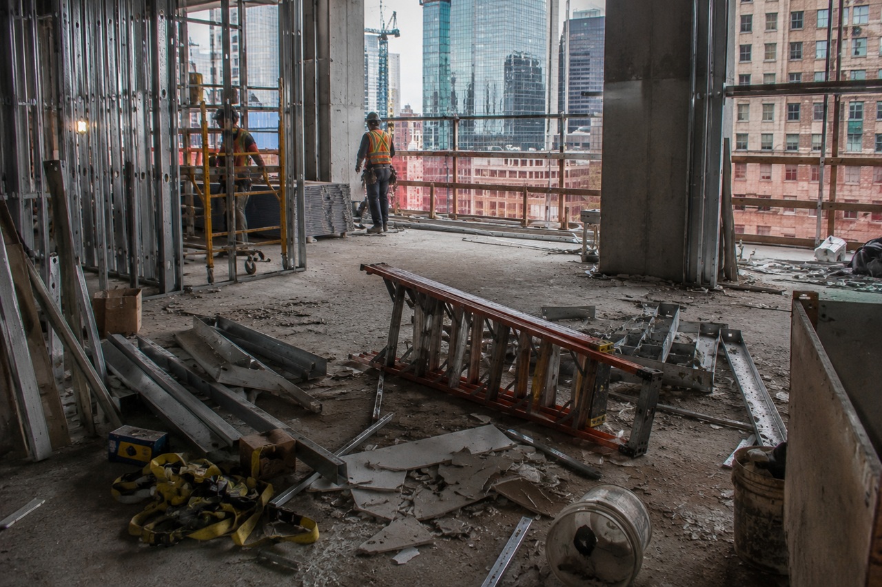 A fallen stepladder lies on its side amidst construction debris, drywall pieces, and metal studs on the dusty upper floor of a New York City high-rise construction site, while two workers in safety vests stand near the open edge overlooking city skyscrapers in the background.